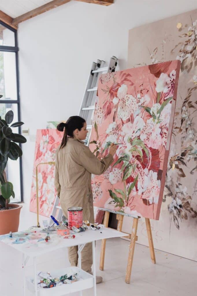 Artist painting floral artwork on canvas in studio, surrounded by vibrant colors and creative tools. Modern art scene.