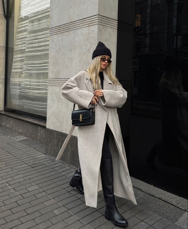 Stylish woman in a gray coat, black boots, and beanie, holding a designer handbag on a city street.