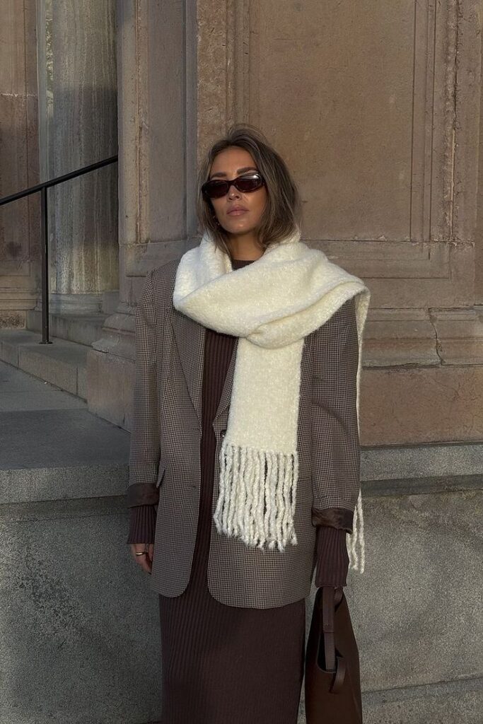 Woman in stylish brown outfit, textured scarf, and sunglasses stands against architectural backdrop.