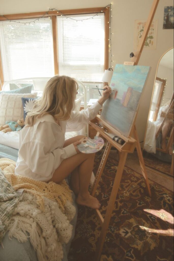 Artist painting on canvas in cozy room, sitting on bed with art supplies and soft lighting, creating a serene creative atmosphere.