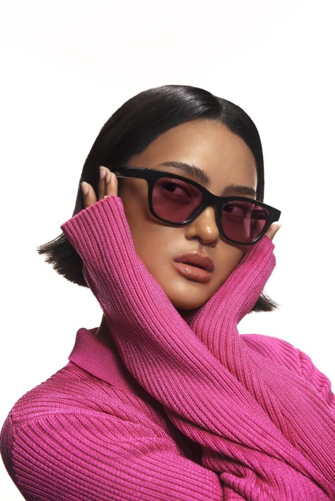 Woman in pink sweater and sunglasses, posing stylishly with hands near face against white background.