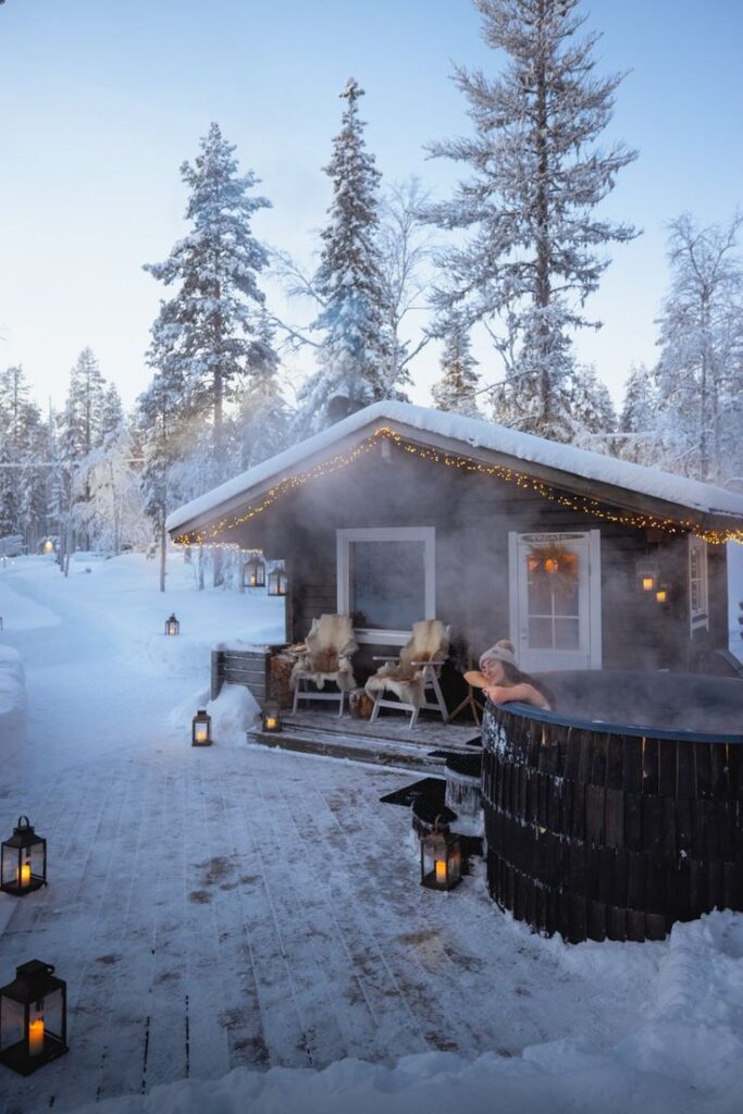 Cozy winter cabin with a person relaxing in a hot tub, surrounded by snowy forest and lanterns.