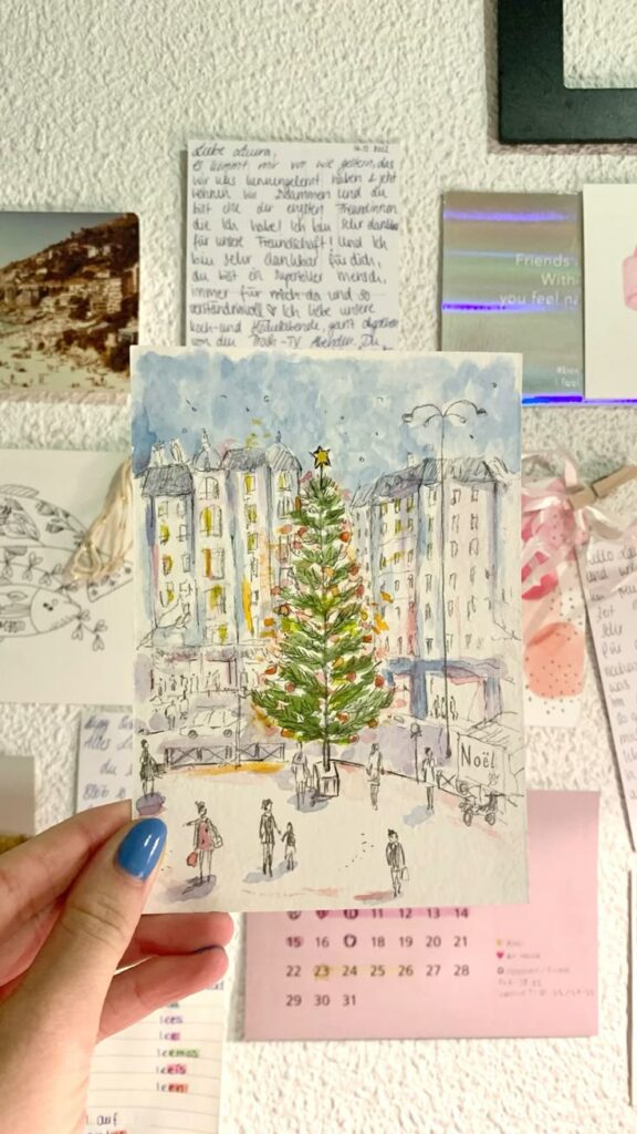 Postcard Christmas ⭐️ Hand holding a festive painting of a Christmas tree in a snowy city, displayed on a photo wall. | Sky Rye Design Hand holding a festive painting of a Christmas tree in a snowy city, displayed on a photo wall.