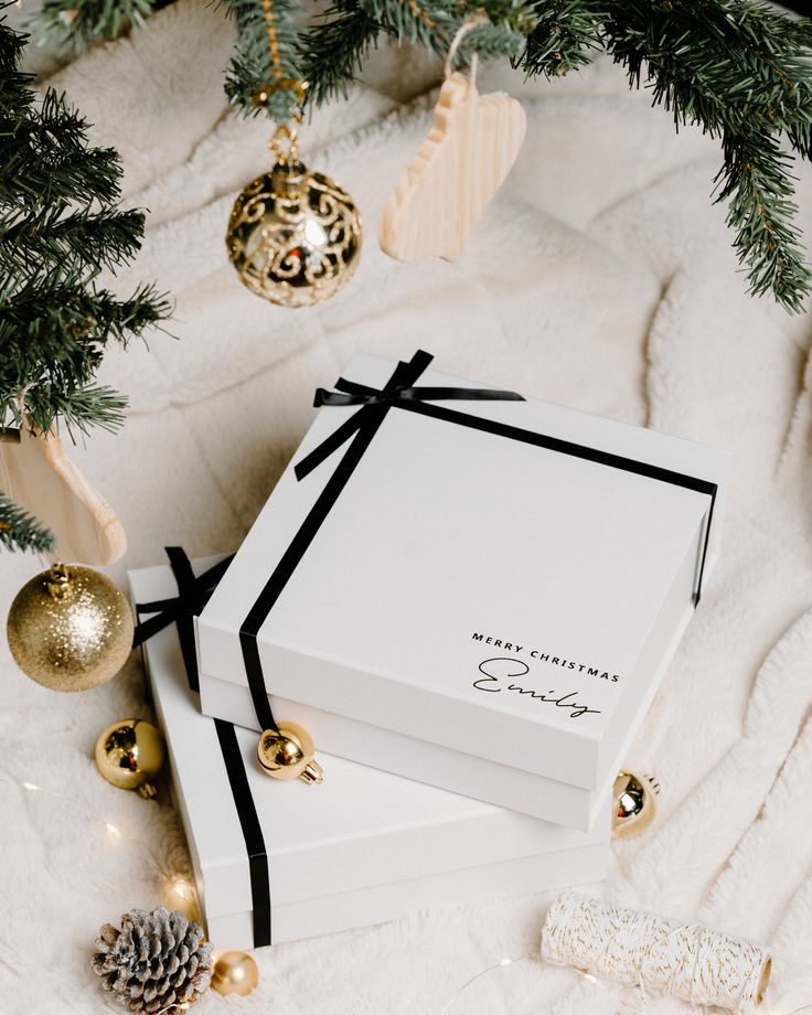 Boîte Co Elegant Christmas gift boxes with black ribbons beneath a decorated tree, featuring gold ornaments and festive pinecones on a cozy blanket. | Sky Rye Design Elegant Christmas gift boxes with black ribbons beneath a decorated tree, featuring gold ornaments and festive pinecones on a cozy blanket.