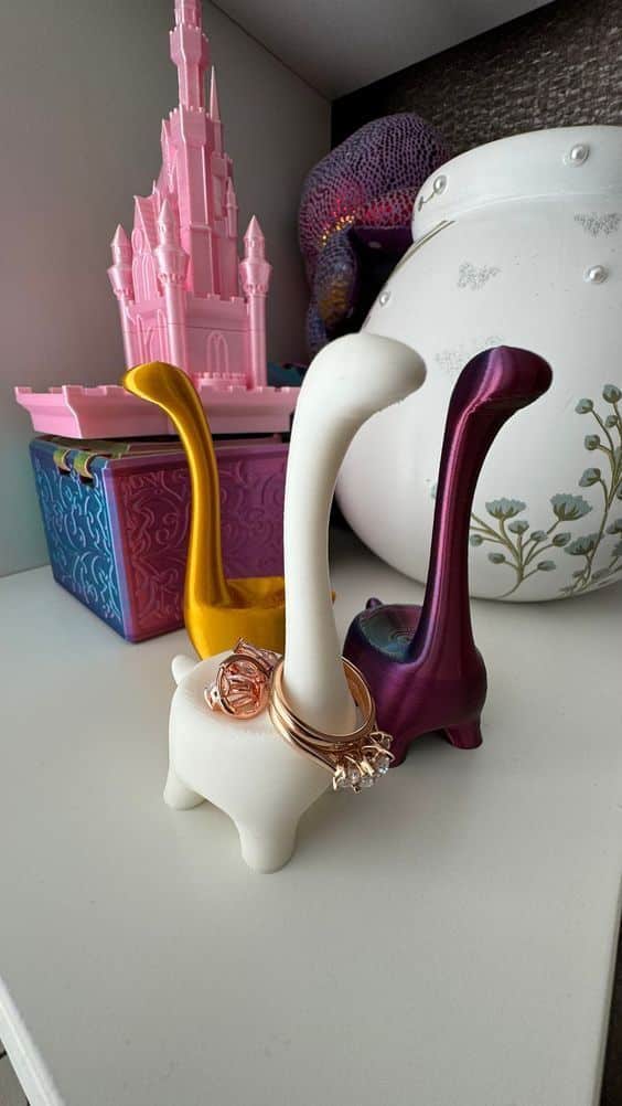 Colorful dinosaur-shaped ring holders with rings displayed on their necks, set beside a pink castle model on a shelf.
