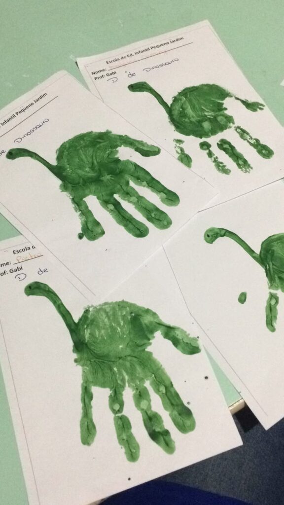 Projeto dinossauros atividade com tinta Green dinosaur handprint art on white paper, showcasing creativity in a classroom setting with children's playful designs. | Sky Rye Design Green dinosaur handprint art on white paper, showcasing creativity in a classroom setting with children's playful designs.