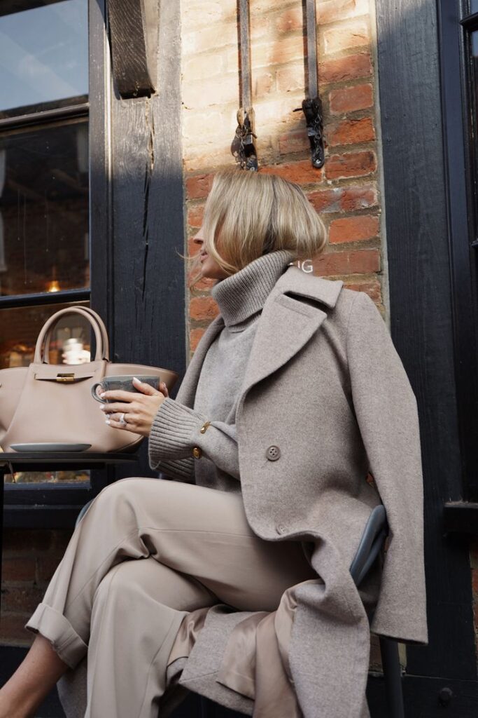 Fashionable woman in beige coat and turtleneck, sitting outdoors at café with handbag and coffee cup. Cozy fall style.