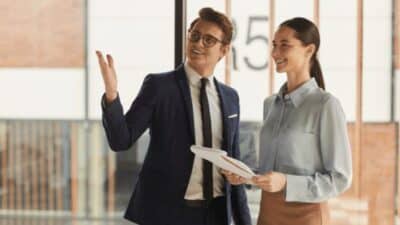 Two professionals discussing business strategies, one holding a tablet, standing in a modern office environment.