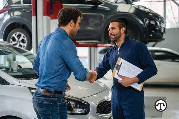 Man greeting a mechanic with a handshake at an auto repair shop Mechanic in blue coveralls shakes hands with customer in garage, car lifted behind, clipboard held. | Sky Rye Design Mechanic in blue coveralls shakes hands with customer in garage, car lifted behind, clipboard held.