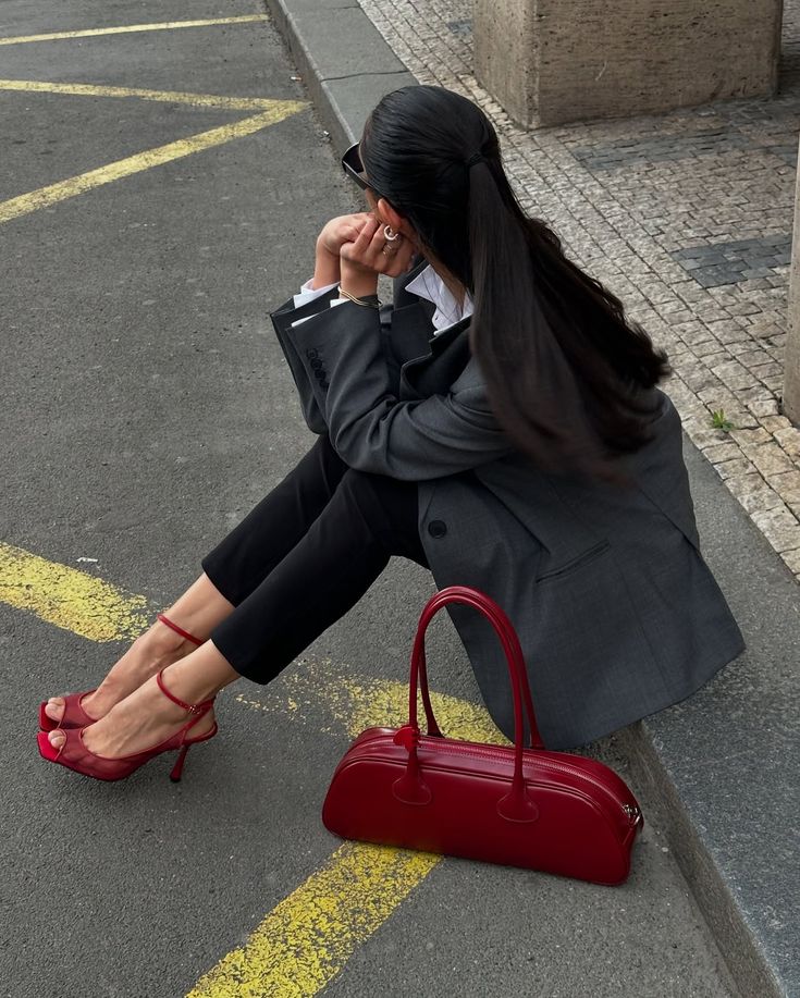 Fashionable woman in gray blazer and red heels sitting on curb, with matching red handbag on street.