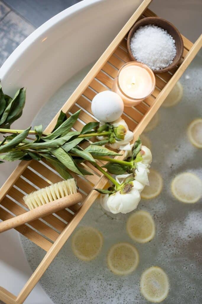 Relaxing bath setup with flowers, candle, bath bomb, brush, and salts on a wooden tray over a tub with lemon slices. Spa essentials.