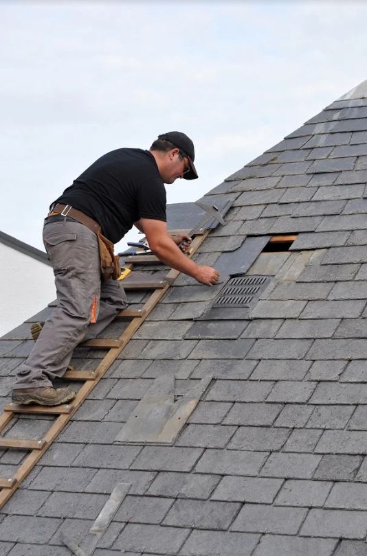 Roofing_ Repair or Replace_ Worker repairing slate shingles on a roof, using tools while standing on a ladder. | Sky Rye Design Worker repairing slate shingles on a roof, using tools while standing on a ladder.