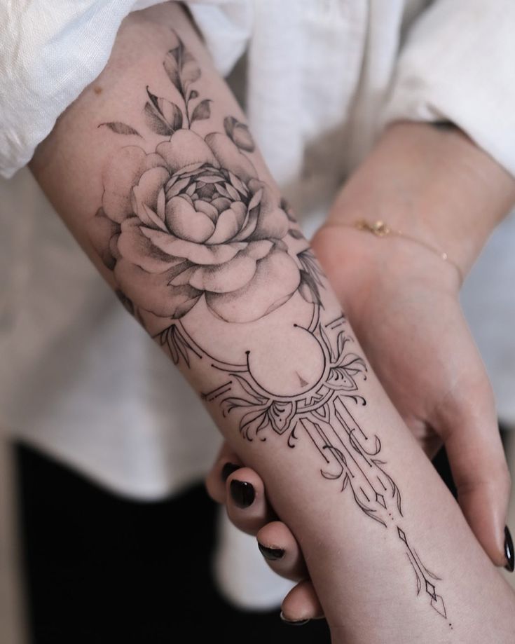 Black and grey floral tattoo on forearm featuring a detailed rose design, showcasing delicate shading and intricate linework.