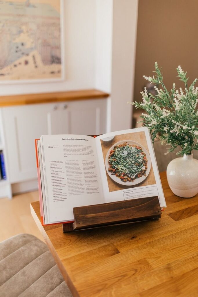 Rustic Wooden Cookbook Stand - Farmhouse Kitchen Book Holder Tablet Stand Recipe Display Handmade Gift Open cookbook displaying a colorful recipe on a wooden stand with a green plant in a vase, set in a cozy kitchen space. | Sky Rye Design Open cookbook displaying a colorful recipe on a wooden stand with a green plant in a vase, set in a cozy kitchen space.