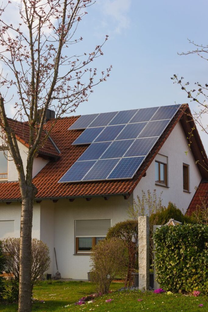 RooftopWithSolarPanelsAndYellowFlowersAtSouthGermany House with rooftop solar panels and red tiles, surrounded by trees under a clear blue sky. | Sky Rye Design House with rooftop solar panels and red tiles, surrounded by trees under a clear blue sky.