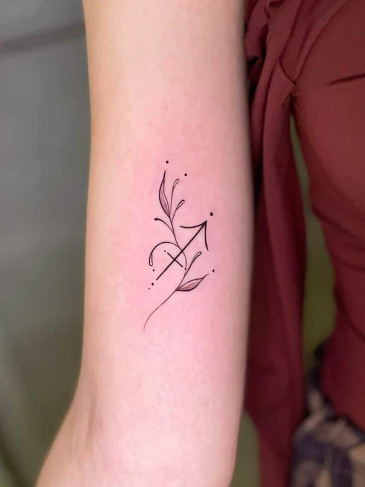 Minimalist black ink arrow tattoo with leaves and dots on upper arm, designed with elegant, flowing lines.