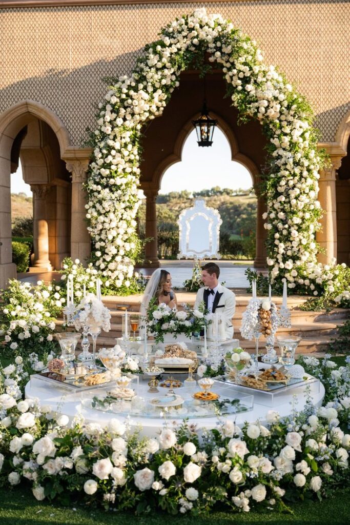 Sarah and David The Besharati Group 1 Elegant wedding setting with a couple seated at a lavish table, adorned with white flowers and a scenic backdrop. | Sky Rye Design Elegant wedding setting with a couple seated at a lavish table, adorned with white flowers and a scenic backdrop.