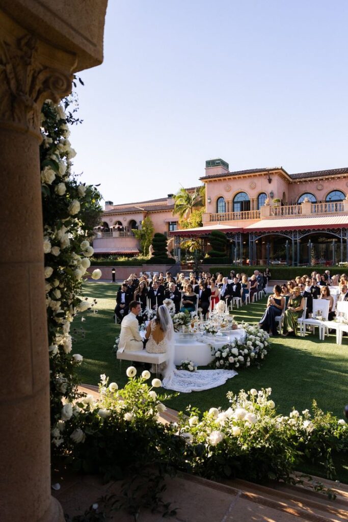 Sarah and David The Besharati Group Outdoor wedding ceremony at a luxurious estate, with a couple seated at a floral-adorned table and guests watching in elegant attire. | Sky Rye Design Outdoor wedding ceremony at a luxurious estate, with a couple seated at a floral-adorned table and guests watching in elegant attire. small wedding