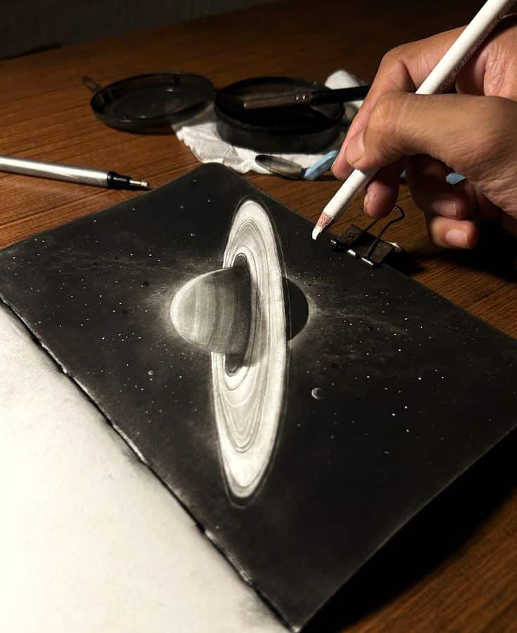 Artist sketching Saturn with rings in pencil on black paper, wood desk background, close-up, art tools nearby, creative space.