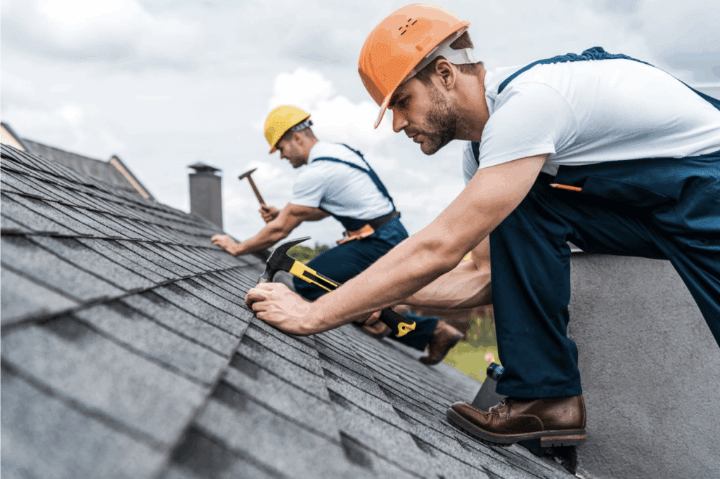 Roofers in hard hats and overalls working on shingled roof with hammers.