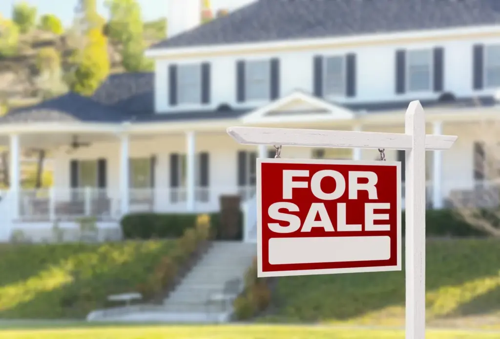 Red For Sale sign in front of a large white house with green lawn, symbolizing a real estate opportunity.
