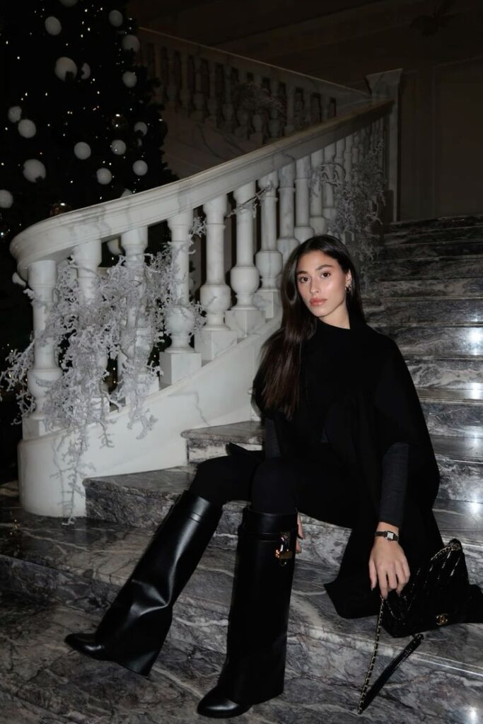 Woman in elegant black outfit and knee-high boots sits on marble stairs, next to festive decor and a Christmas tree.