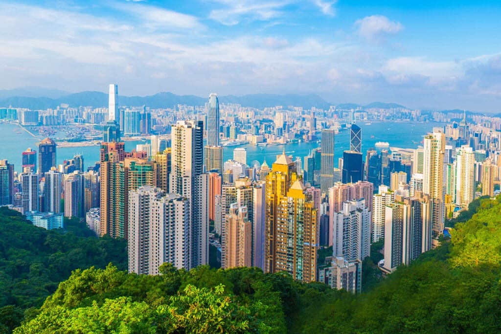 Skyline view of Hong Kong with tall skyscrapers, lush greenery, and Victoria Harbour under a blue sky. Perfect blend of nature and cityscape.