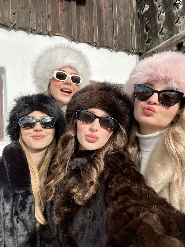 Four women in stylish fur coats and sunglasses pose for a selfie outdoors in winter.