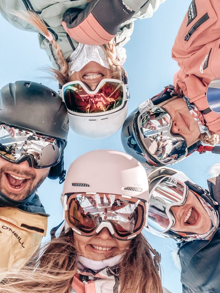 Group of friends smiling in ski gear, looking down at the camera on a sunny day, enjoying snowy mountain adventure.