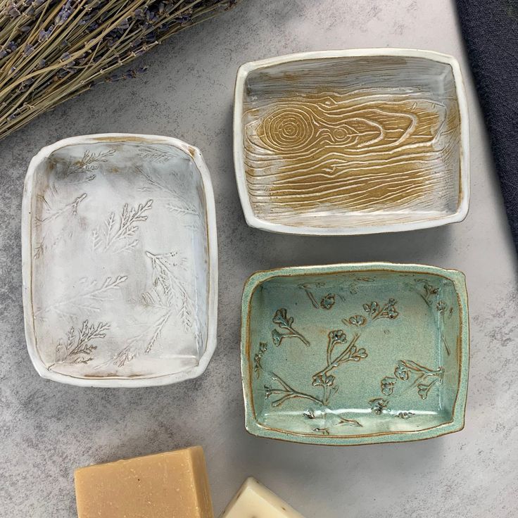 Three handcrafted ceramic soap dishes with nature-inspired designs on a textured grey background, next to bars of soap and dried lavender.