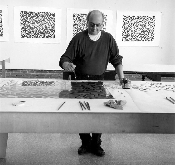 Artist working on abstract designs in a studio, surrounded by intricate pattern artworks and art tools on a large table.