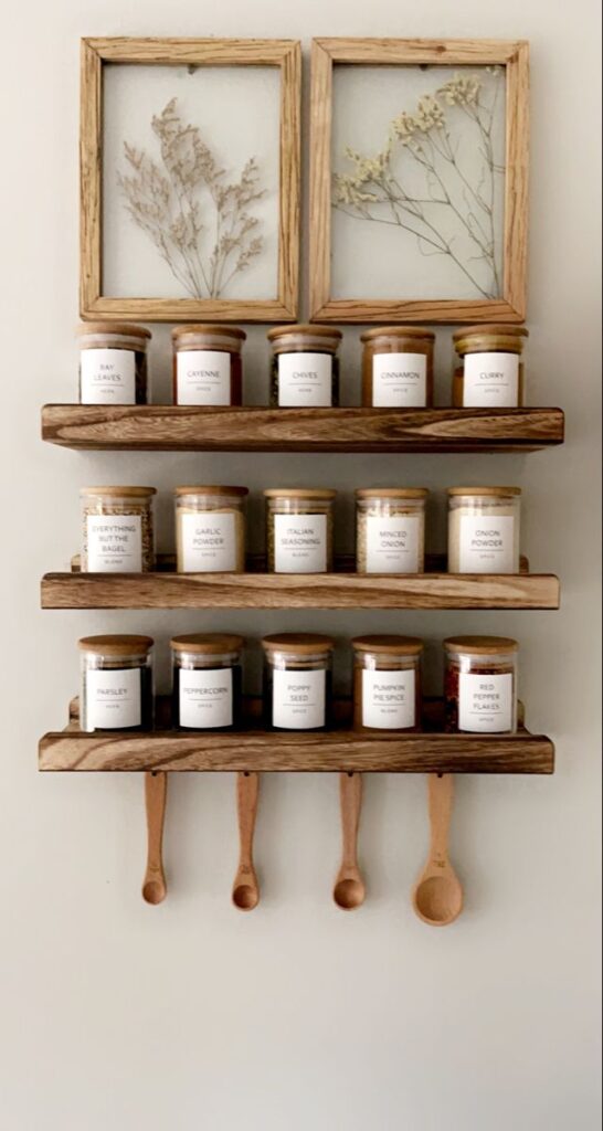 Wooden spice rack with labeled jars and spoons, featuring herbs and spices like bay leaves and cinnamon, and framed dried plants above.