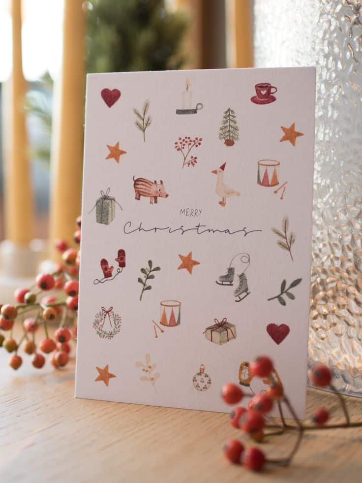 Christmas card with festive illustrations, including a candle, tree, and gifts, surrounded by seasonal decorations on a wooden table.