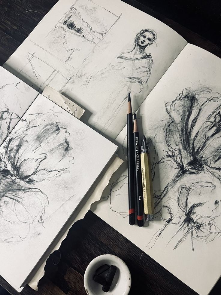 Sketchbooks with charcoal drawings of flowers and a woman, accompanied by pencils on a wooden table.