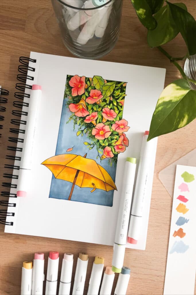 Colorful drawing of flowers and umbrella on sketchbook surrounded by marker pens on wooden table.