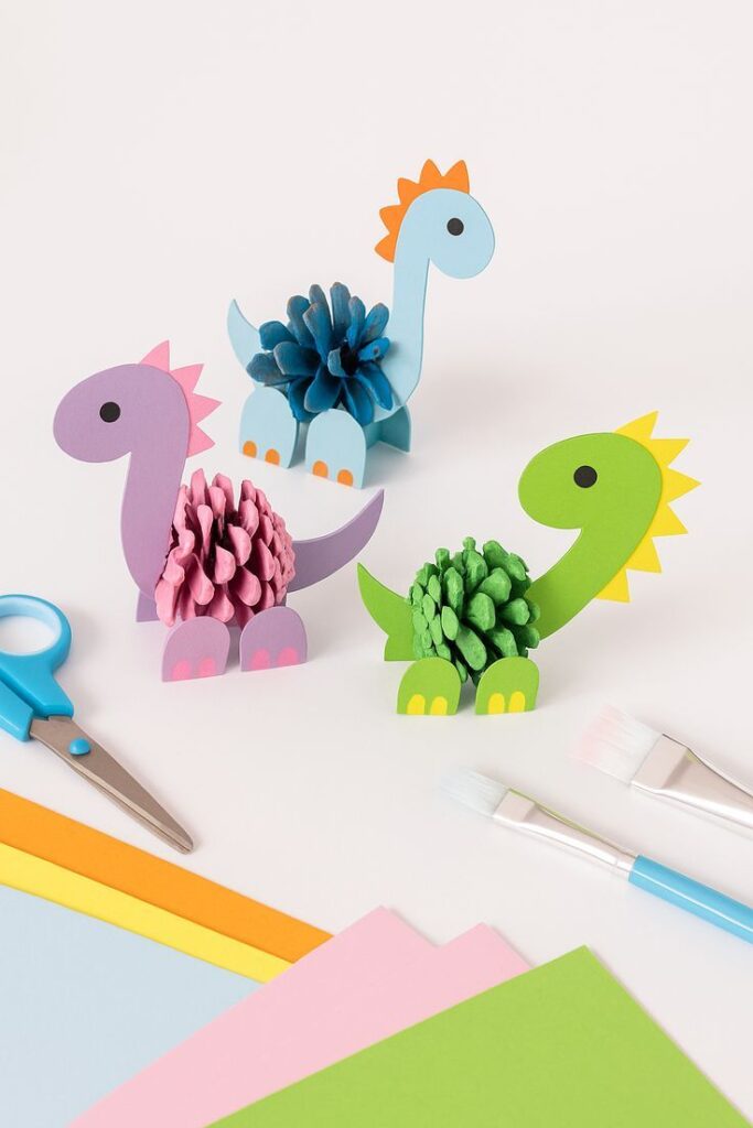Sweet DIY Pinecone Paper Dinosaur Craft for Kids Colorful pinecone dinosaurs craft with blue, pink, and green designs. Scissors, paper, and brushes complete the DIY activity setup. | Sky Rye Design Colorful pinecone dinosaurs craft with blue, pink, and green designs. Scissors, paper, and brushes complete the DIY activity setup.