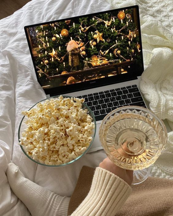 Laptop streaming a Christmas movie with a cozy setup of popcorn and a drink on a bed.