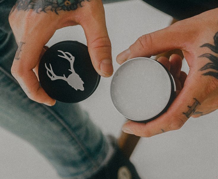 Tattooed hands holding an open hair pomade tin with a stag logo, perfect for grooming and styling. Casual jeans visible in the background.