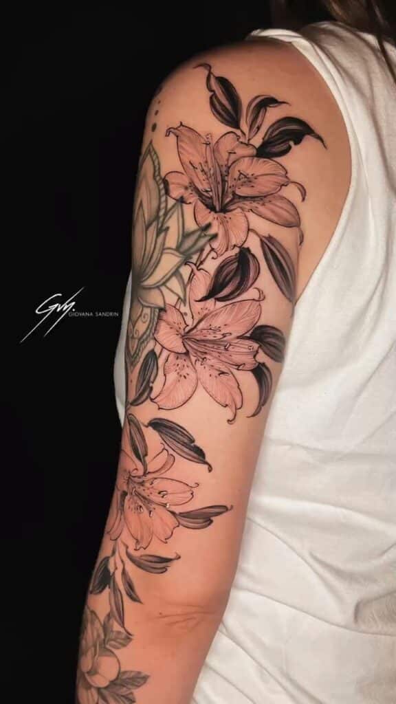 Tatuagem de Lírios _ Tattoo Feminina l Por Giovana Sandrin Arm with a floral tattoo featuring detailed lily designs in soft pink and black ink, extending from shoulder to elbow. | Sky Rye Design Arm with a floral tattoo featuring detailed lily designs in soft pink and black ink, extending from shoulder to elbow.