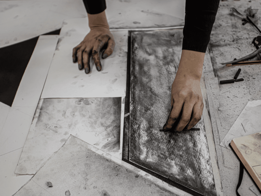 Artist's hands creating a charcoal drawing on paper, surrounded by art tools and sketchbooks.