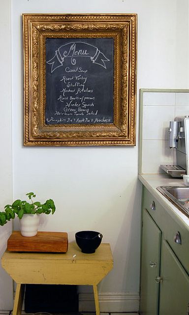 Chalkboard menu in ornate frame lists dishes like carrot soup and roast turkey in vintage kitchen with basil plant on yellow table.