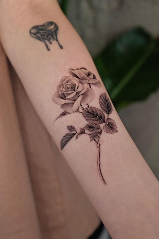 Realistic rose tattoo on forearm with detailed leaves; black ink design beside small heart. Minimalist floral body art.
