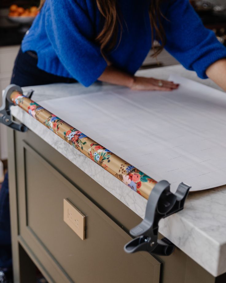 The Art of Holiday Gift Wrapping - Chris Loves Julia Woman wrapping gift on marble countertop using floral paper and plastic clamps for support. Gift wrapping station setup. | Sky Rye Design Woman wrapping gift on marble countertop using floral paper and plastic clamps for support. Gift wrapping station setup.