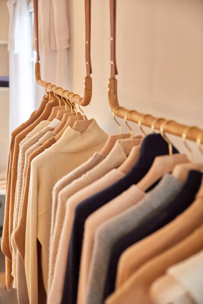 Row of beige and neutral sweaters on wooden hangers in a bright, minimalist closet setting. Cozy, organized, and stylish wardrobe display.
