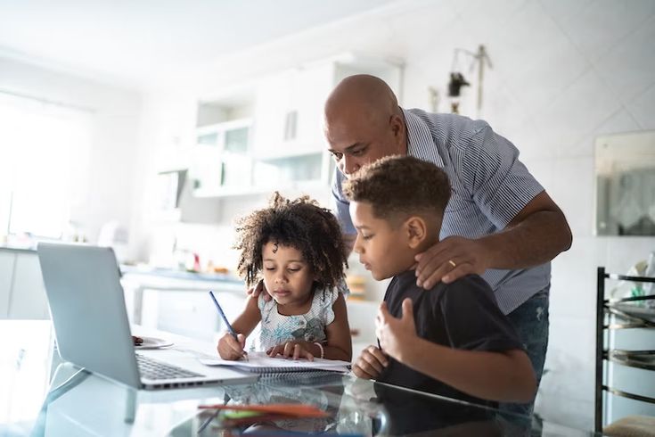 Father helps children with homework on laptop in kitchen, creating a supportive learning environment, enhancing family education moments.