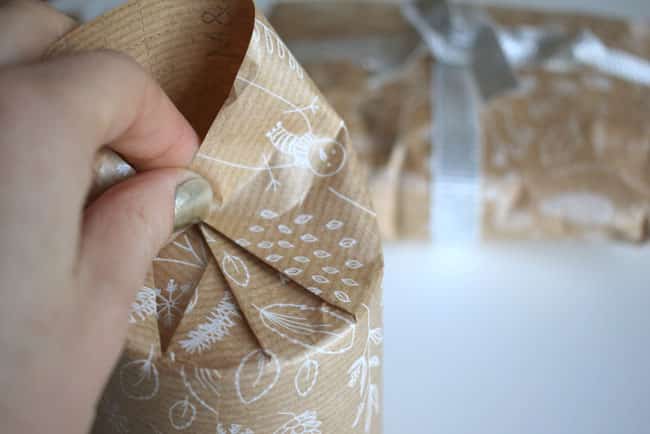 The-pleat-method Hand wrapping a gift with brown paper featuring white holiday patterns. Another wrapped present with a ribbon in the background. | Sky Rye Design Hand wrapping a gift with brown paper featuring white holiday patterns. Another wrapped present with a ribbon in the background.