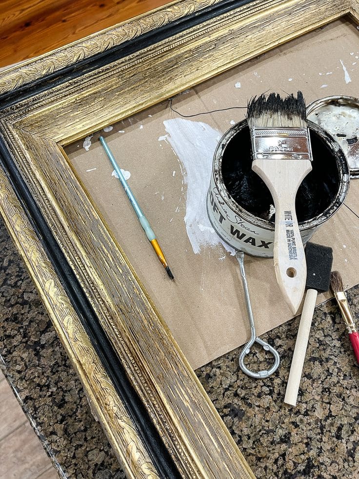 Gold-painted picture frame with paintbrushes, wax, and tools on a work surface, illustrating a DIY frame restoration project.