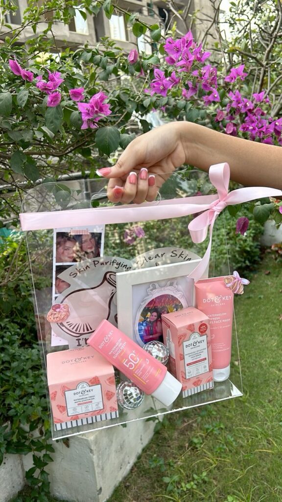 Title_ Adorable Pink Aesthetic Skincare Hamper Hand holding a clear bag with Dot & Key skincare products, decorated with pink flowers in a lush garden setting. | Sky Rye Design Hand holding a clear bag with Dot & Key skincare products, decorated with pink flowers in a lush garden setting.