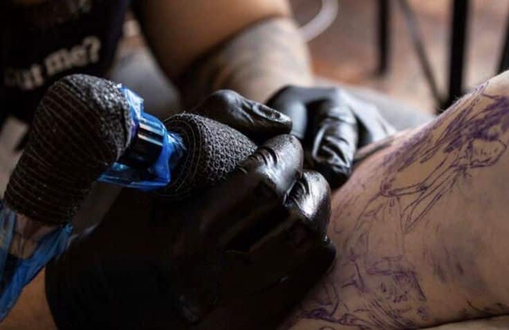 Tattoo artist with black gloves working on detailed leg tattoo design using a tattoo machine.