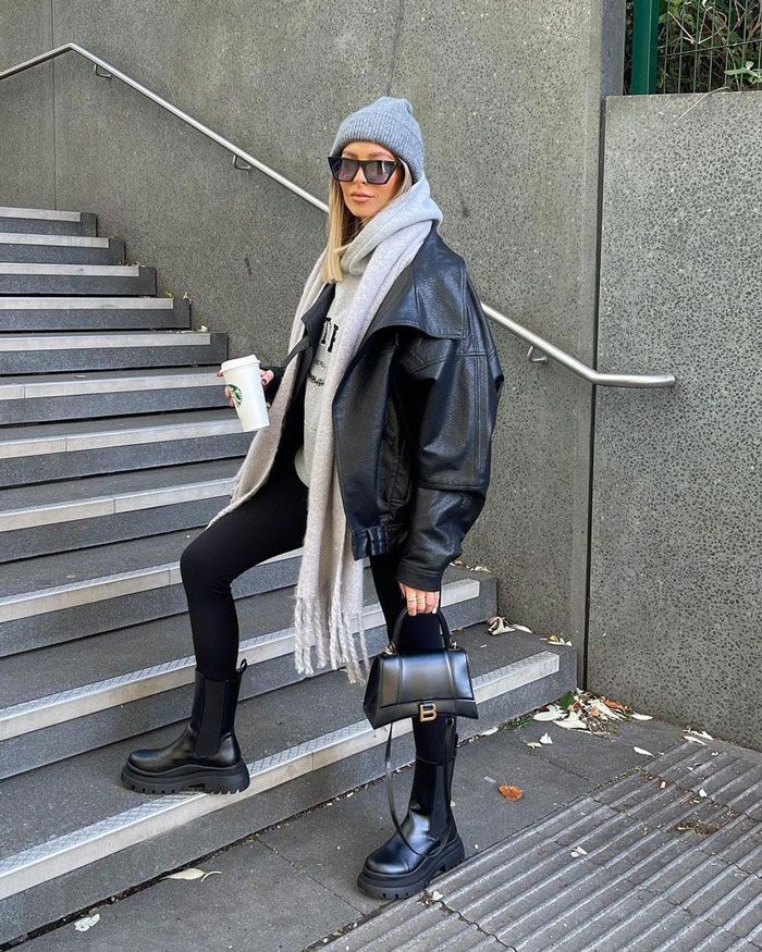 Stylish woman in winter outfit with leather jacket, beanie, and boots, holding coffee on stairs. Fashionable urban look.
